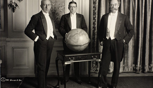 black and white of shackleton amundsen perry in formal dinner jackets around large globe on display table