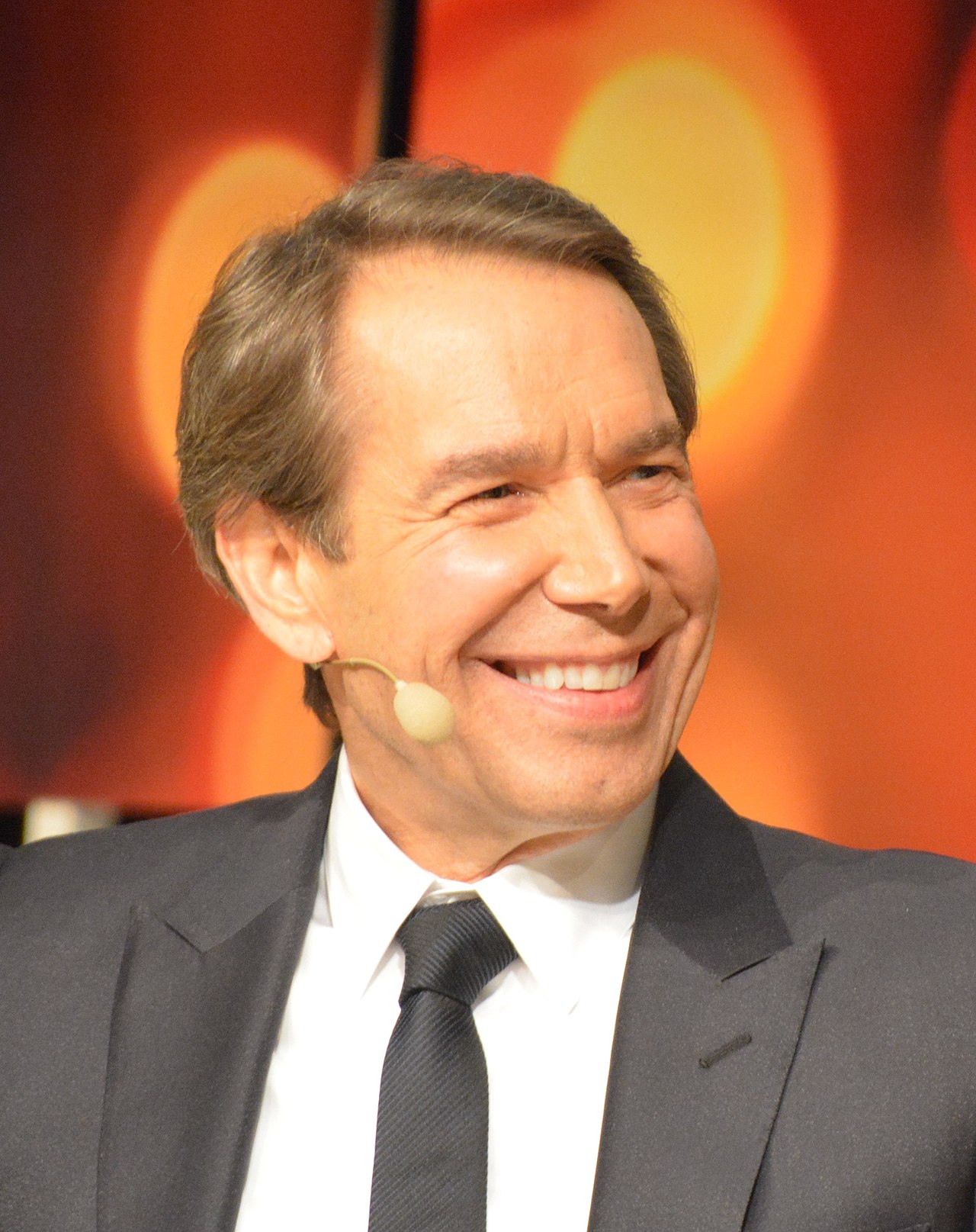 jeff koons color smiling headshot great suit tie microphone orange background