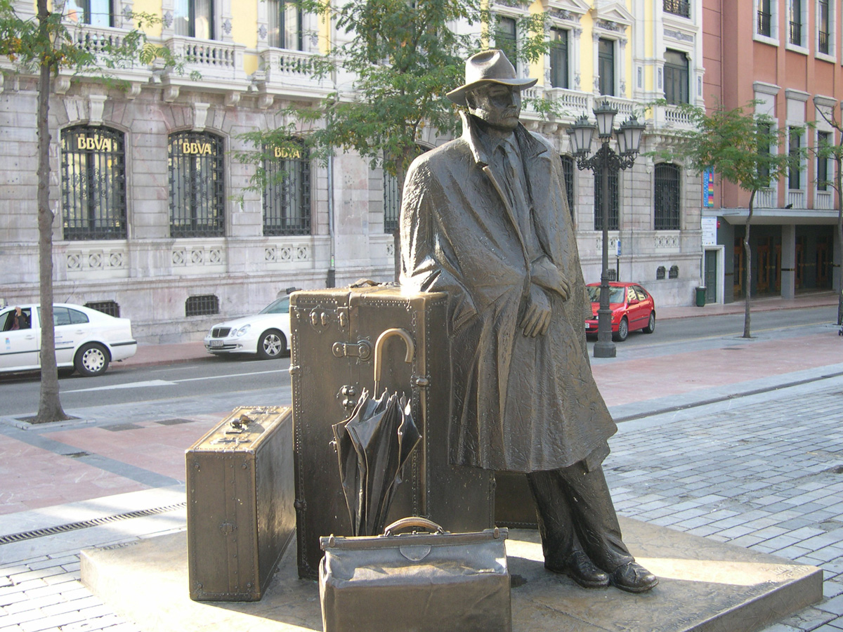 traveler statue on street oviedo spain man in overcoat and hat with umbrella and briefcase and suitcase @servetolead.com